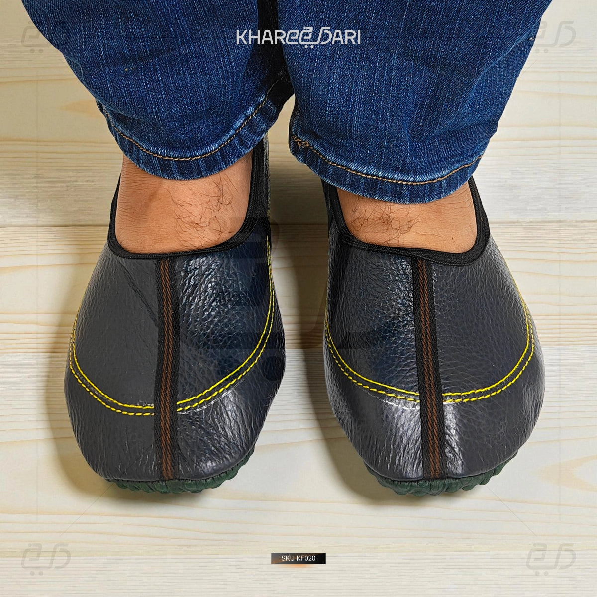 Black leather slippers with yellow stitching worn by a person in blue jeans on a light wooden floor. Purple