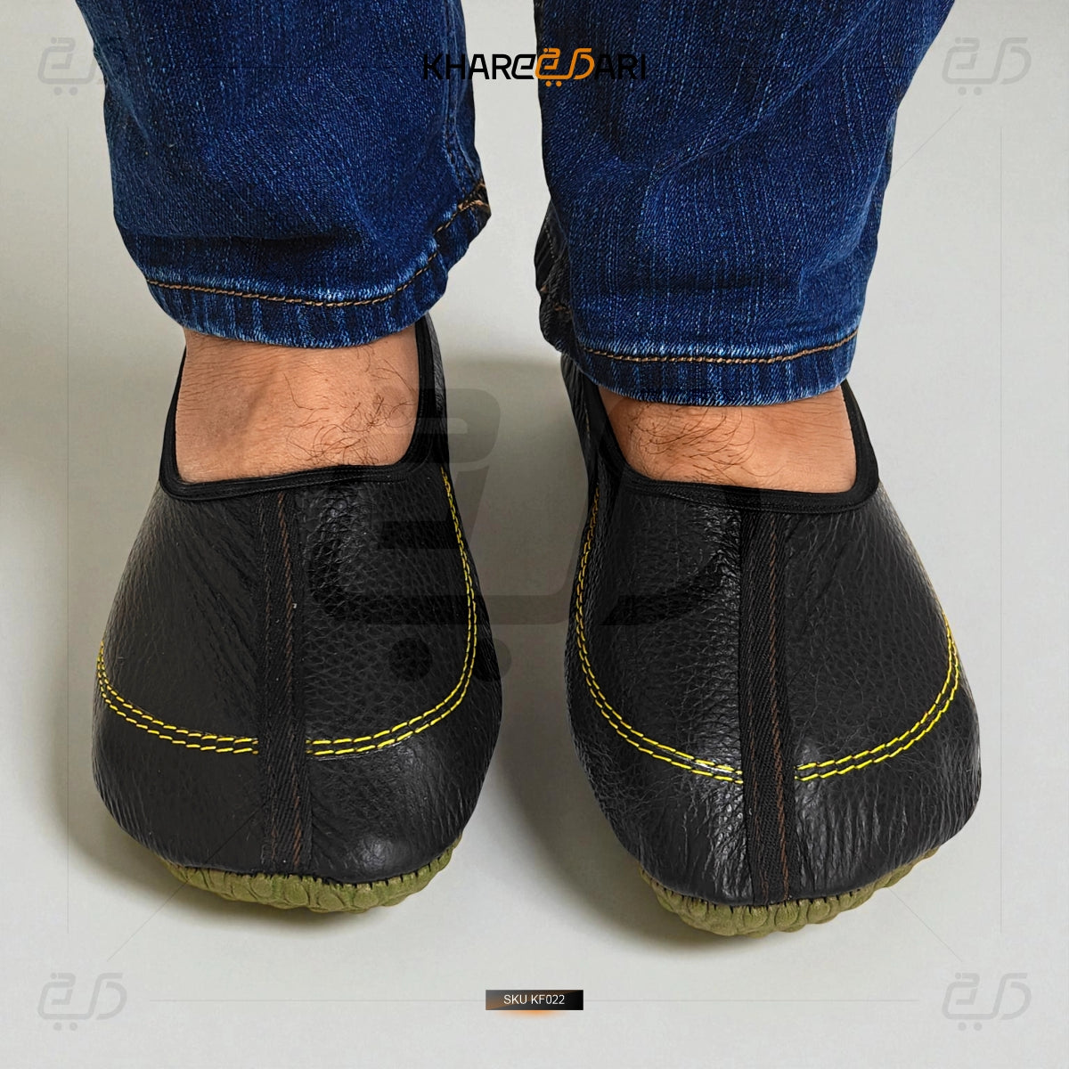 Black leather slippers with yellow stitching worn with blue jeans on a light gray background. Black